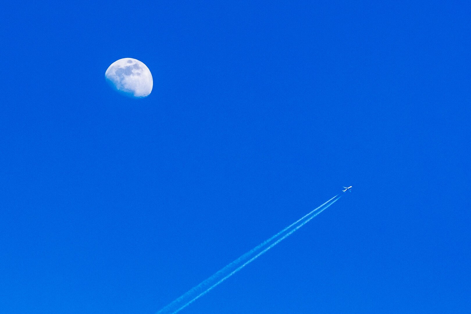 a lone airliner (contrails visible) cruising by the moon in a cloudless blue sky during daytime