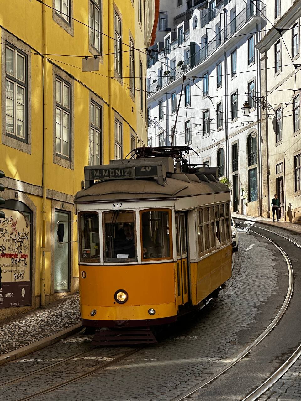 A yellow tram car coming down a hill with a yellow building in the left and white buildings on the right