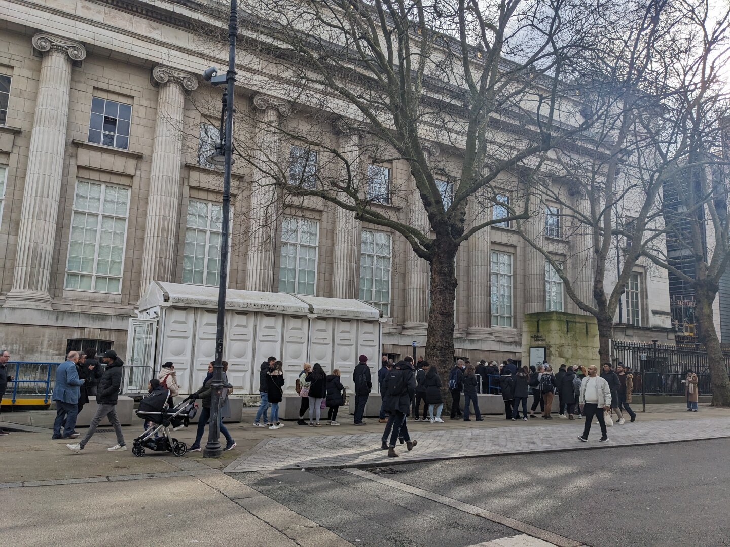 A large pillared building with a big tree and a queue of people in front of it