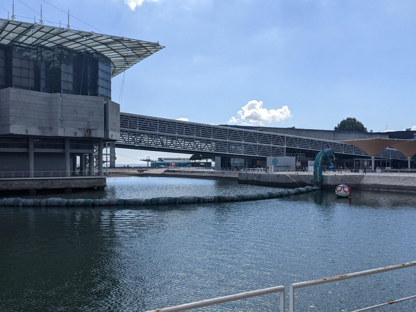 A modern looking building surrounded by water with a covered walkway to the mainland. A large sea monster is in front of the building