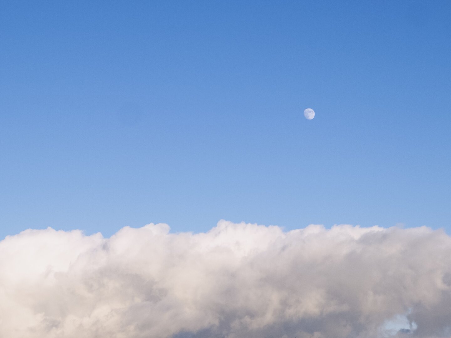 Der Mond über einer Wolkenschicht. Dahinter der blaue Himmel.