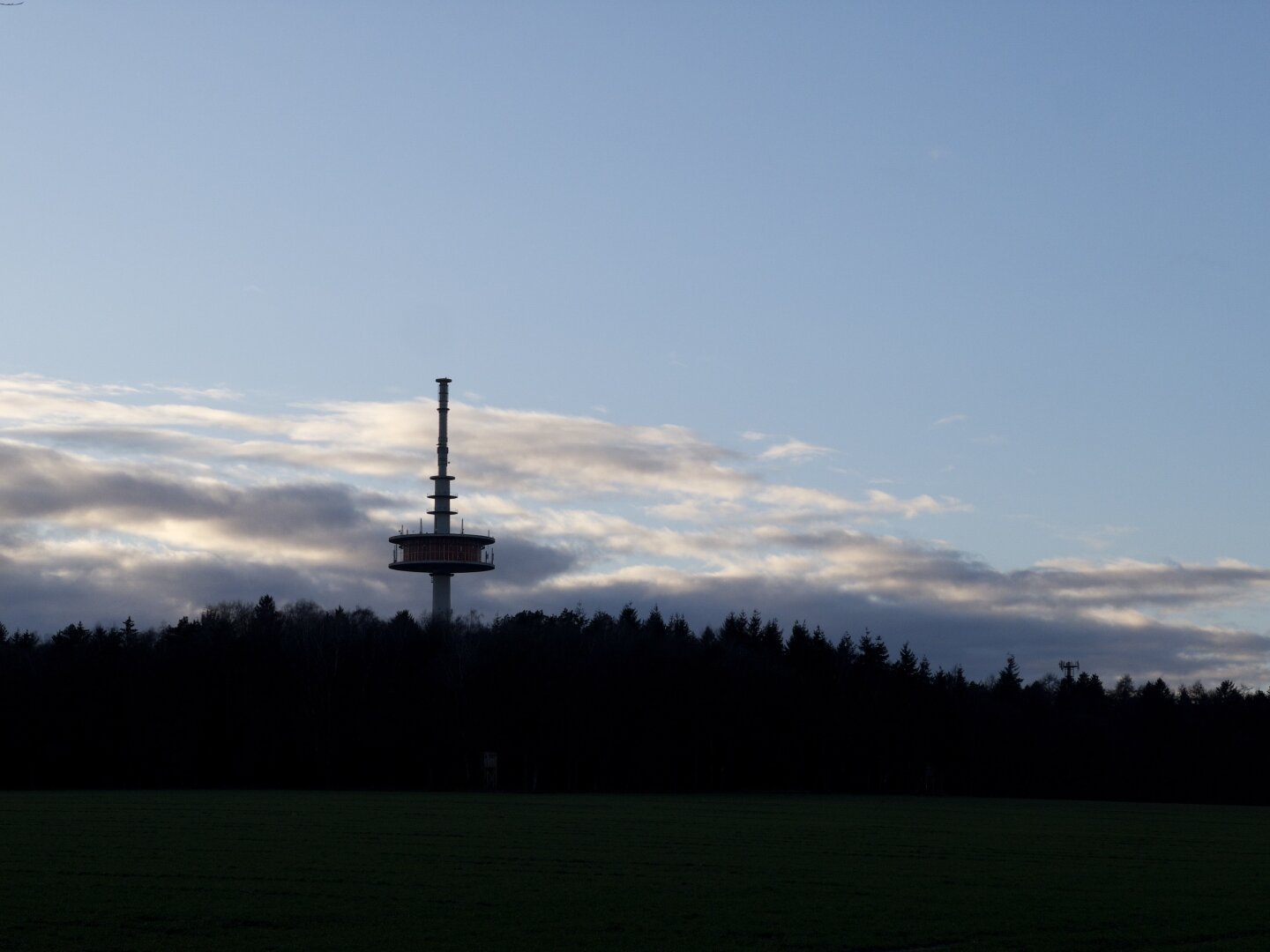 Ein Fernsehturm vor dem Himmel, im Gegenlicht. Der Turm und der Wald davor sind fast nur als Silhouette zur erkennen.