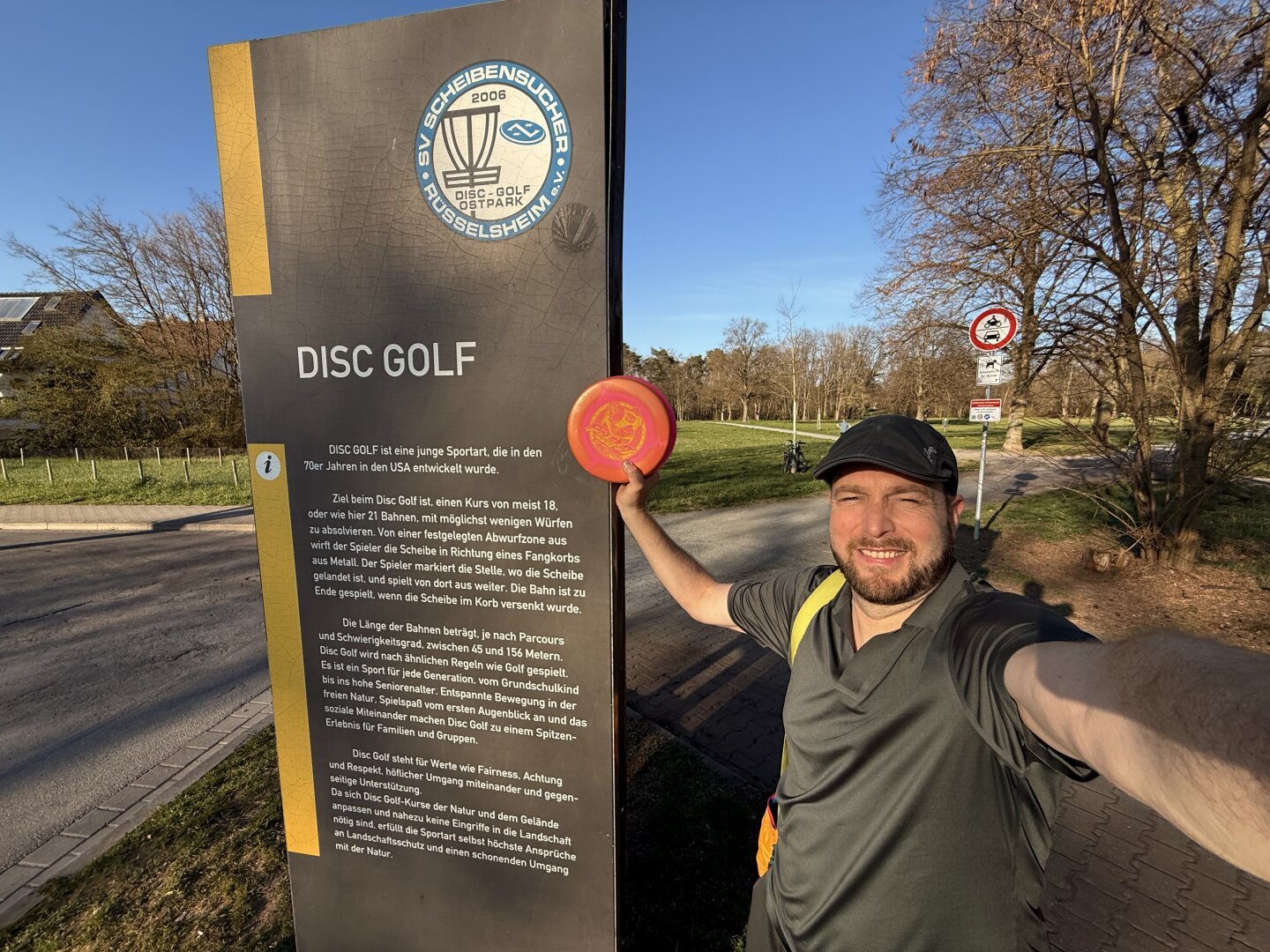 Wolf holding his Kat Mertsch #Polecat in front of the Rüsselsheim 24 holes disc golf course sign outside the park.