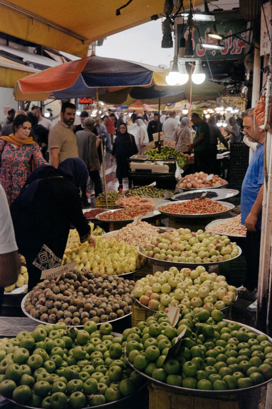 A busy outdoor market with large bowls of colorful fruits like green apples, figs, and pears. People shop under colorful umbrellas and warm hanging lights.