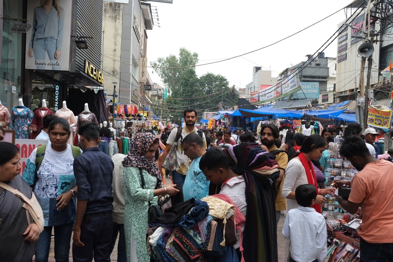 A busy street market with various stalls selling clothes and accessories. Many people are shopping and browsing through the items, including women in traditional attire. Mannequins displaying dresses are visible in front of a shop, and multiple shop signs can be seen in the background.