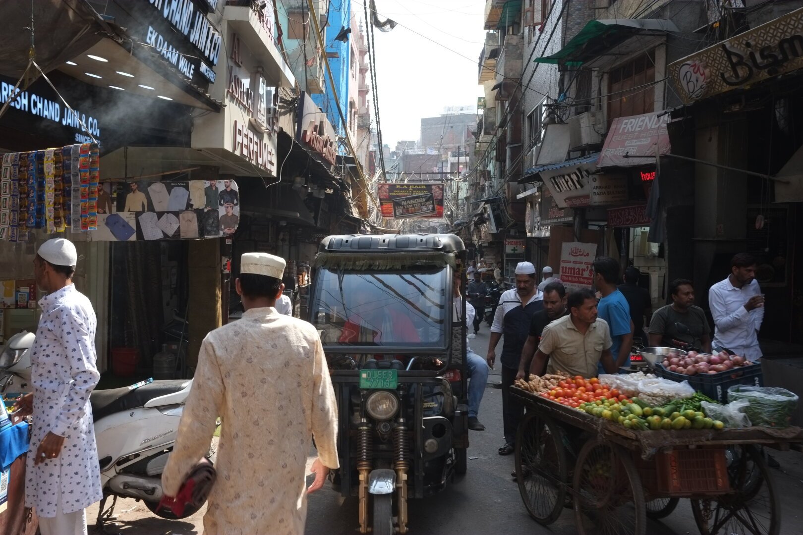 A busy market street with narrow roads, crowded with people in traditional attire, a three-wheeled vehicle, and street vendors selling produce. The area is lined with closely packed shops and signs.