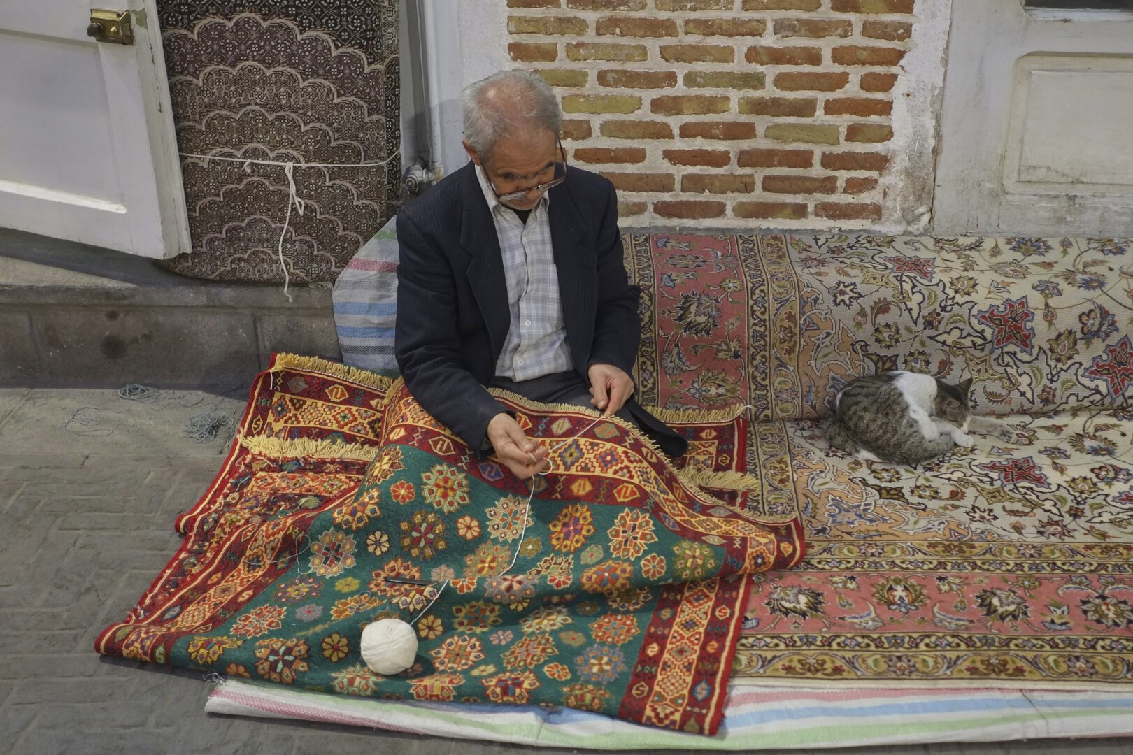 An elderly man sewing a colorful rug, with a ball of white yarn resting on the rug. The man is sitting on another rug on the ground, and a cat is sleeping beside him on the same rug.