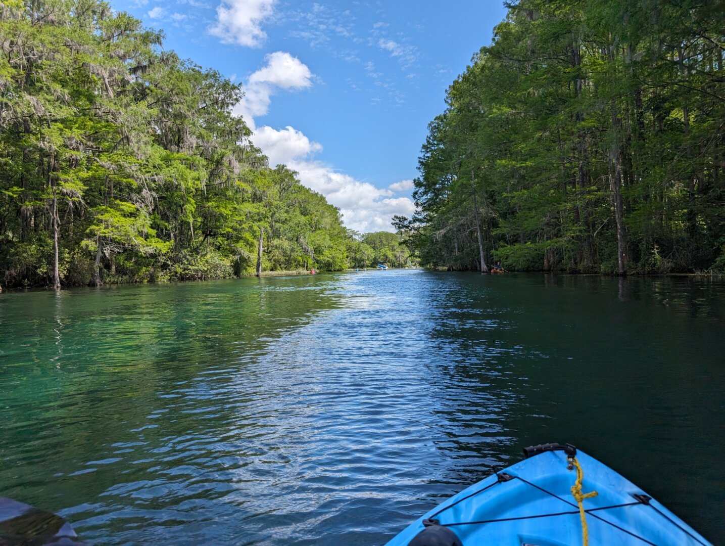 The view from a blue kayak on a bright blue-green river with vibrant green trees on either side and a beautiful, mostly clear sky