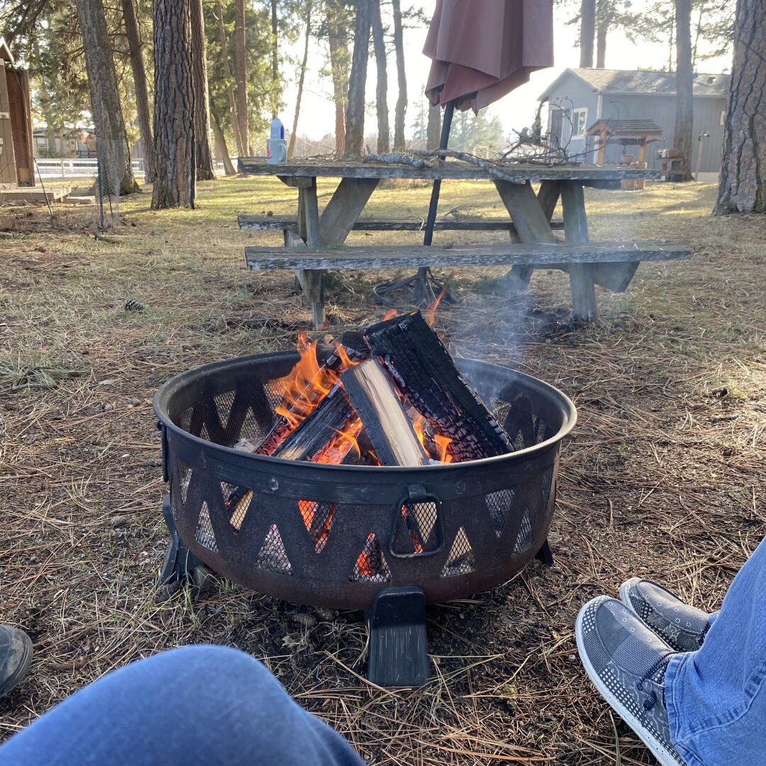 Metal fire pit with a large flame inside. Picnic table with umbrella in background and behind that a small house.