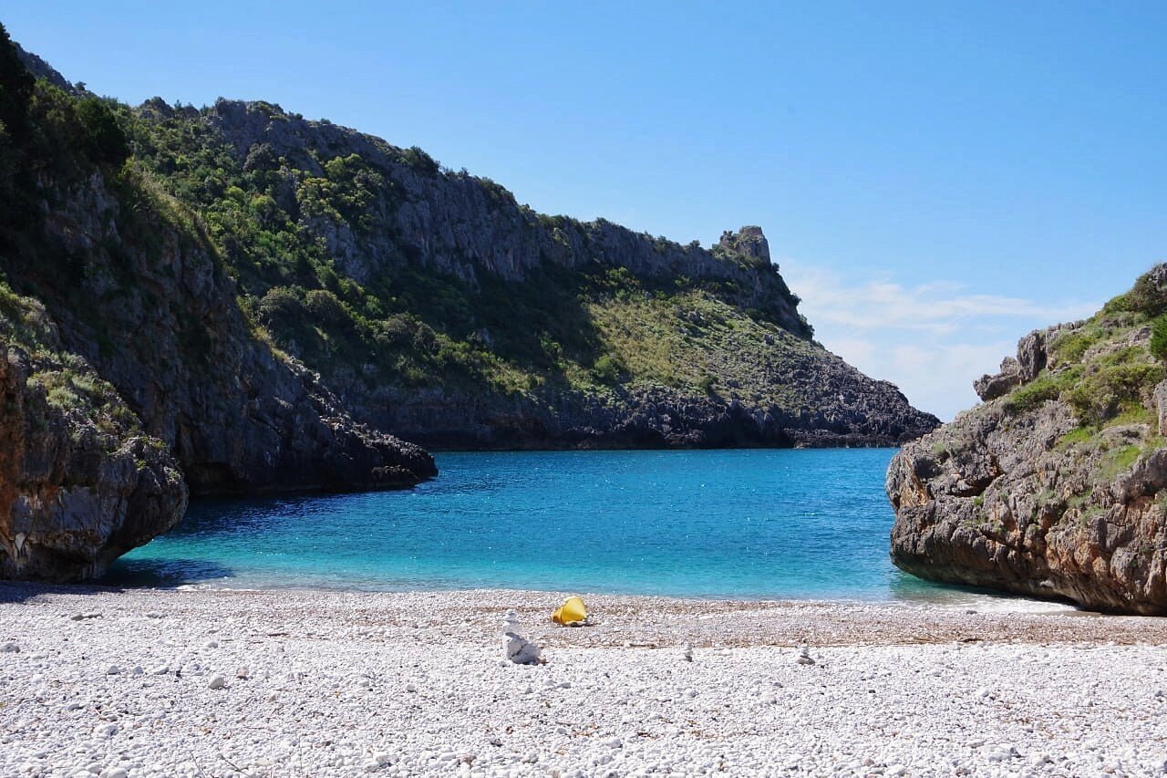 Small white pebble beach nestled in a rocky cove. A Saracen watchtower is visible in the background. The seawater is crystal clear. A yellow buoy and a pile of white stones are in the center of the beach.