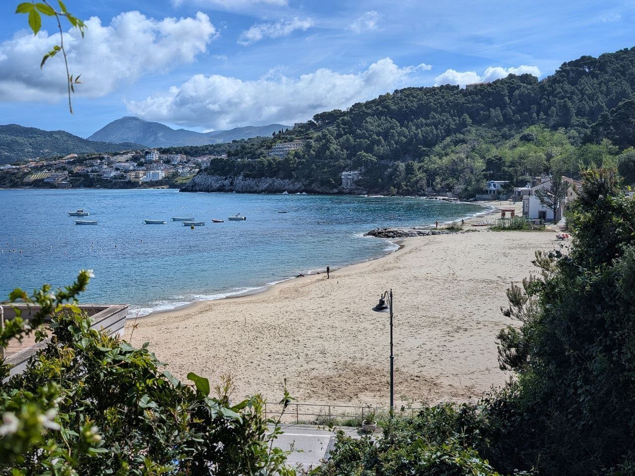 Beach near the port of Palinuro. In the water there are a few small boats. In the background, the village of Palinuro. A person is observing the sea from the beach.