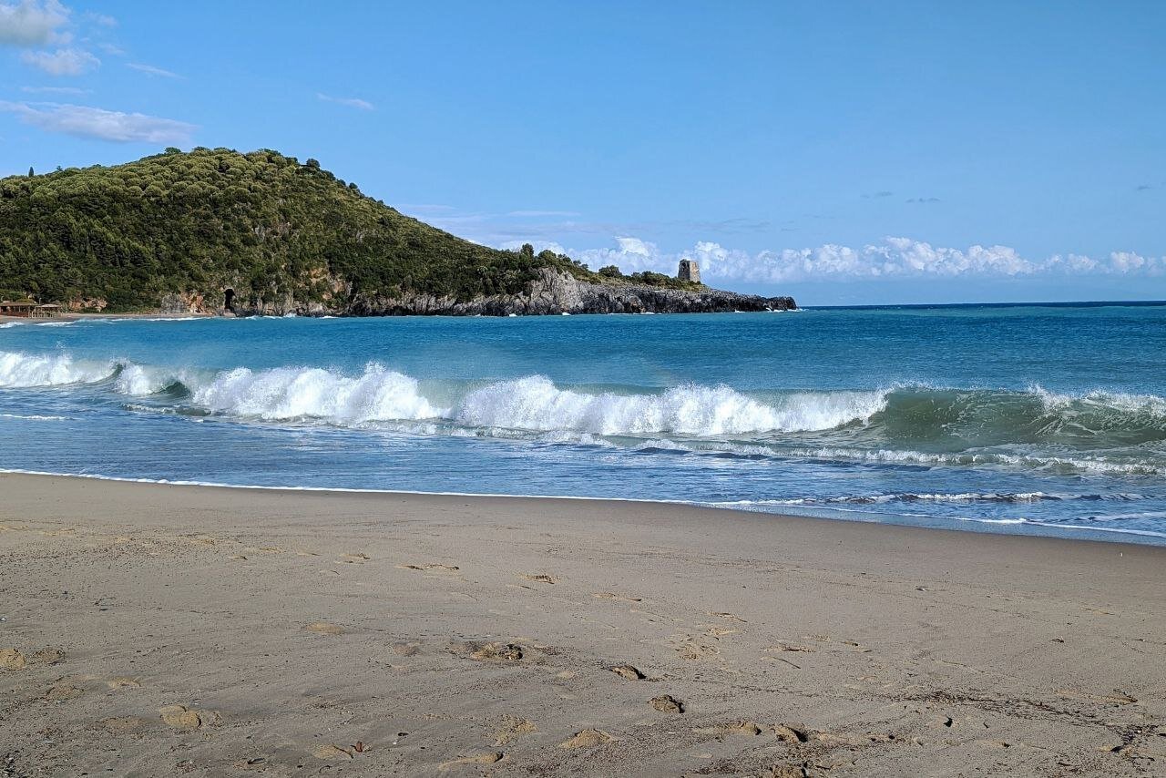 A wave crashes on the beach. In the background, a Saracen tower (Torre dello Zancale) can be glimpsed along the coast.