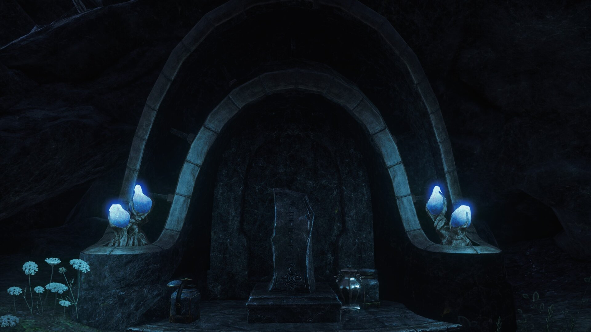 A screenshot from the game Elder Scrolls Online. It depicts the entrance of an Ancestor Tomb, located in Vvardenfell. The entrance is carved into the surrounding rock. It consists of a stone arch which surrounds a stone door. The door is shaped like the arch. There is a stone slab in front of the door, which is shaped like a rectangle, but with jagged edges. Left and right of the door are small planters, which are extensions of the stone arch. They contain blue, fluorescent funghi. The atmosphere is calm and a little bit eerie.