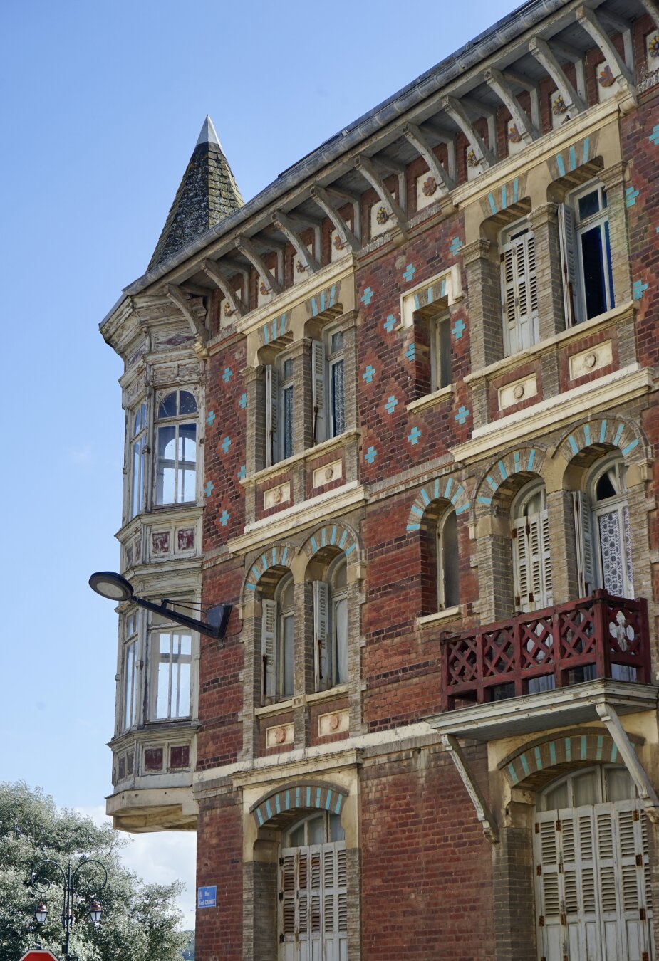 Portion de façade d’une maison d’angle vue de trois quarts. Elle est en briques rouges avec des motifs géométriques ou des rayures selon les étages, et des briques couleur turquoise formant comme des mouchetures. De jolis encadrements de fenêtres en briques et pierre grise rehaussés de bleu turquoise font contraste. Il y a un petit balcon bordé de bois peint en rouge devant une séries de fenêtres sur la droite. A l’angle, un oriel en bois et vitré sur deux niveaux se terminant par une tourelle vient parachever la façade. Il est dans son jus mais très élégant.