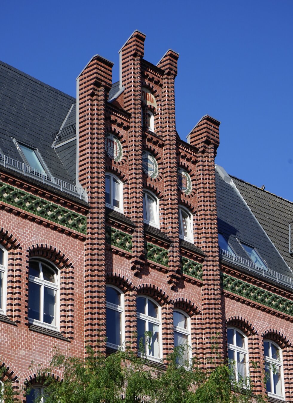 Vue de trois quarts et en contreplongée d’un pignon très pimpant de maison en brique rouge tirant sur le rose. Par endroit les briques rouges alternent avec des briques noires vernissées pour faire joli, il y a également des motifs verts ornementaux. Le pignon se détache d’un bête toit plus moderne, il est en escalier avec une partie centrale plus haute qui porte la date de construction de l’immeuble, 1906. En-dessous, trois motifs circulaires représentent respectivement les armes de Flensbourg, celles de Hambourg, ainsi qu’une poignée de mains.