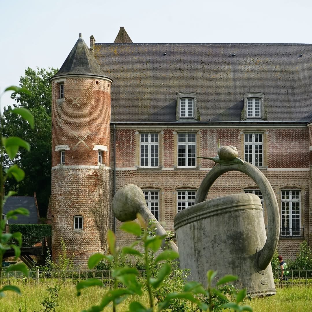 Photo montrant une vue partielle en arrière plan d'un majestueux château en brique avec des grandes tours d'angle.
Manifestement, à certains endroits, les briques sont plus récentes qu'à d'autres. La photo est prise depuis le jardin du château, où trône un immense arrosoir métallique surmonté d'un escargot (il s'agit d'une sculpture-installation artistique).