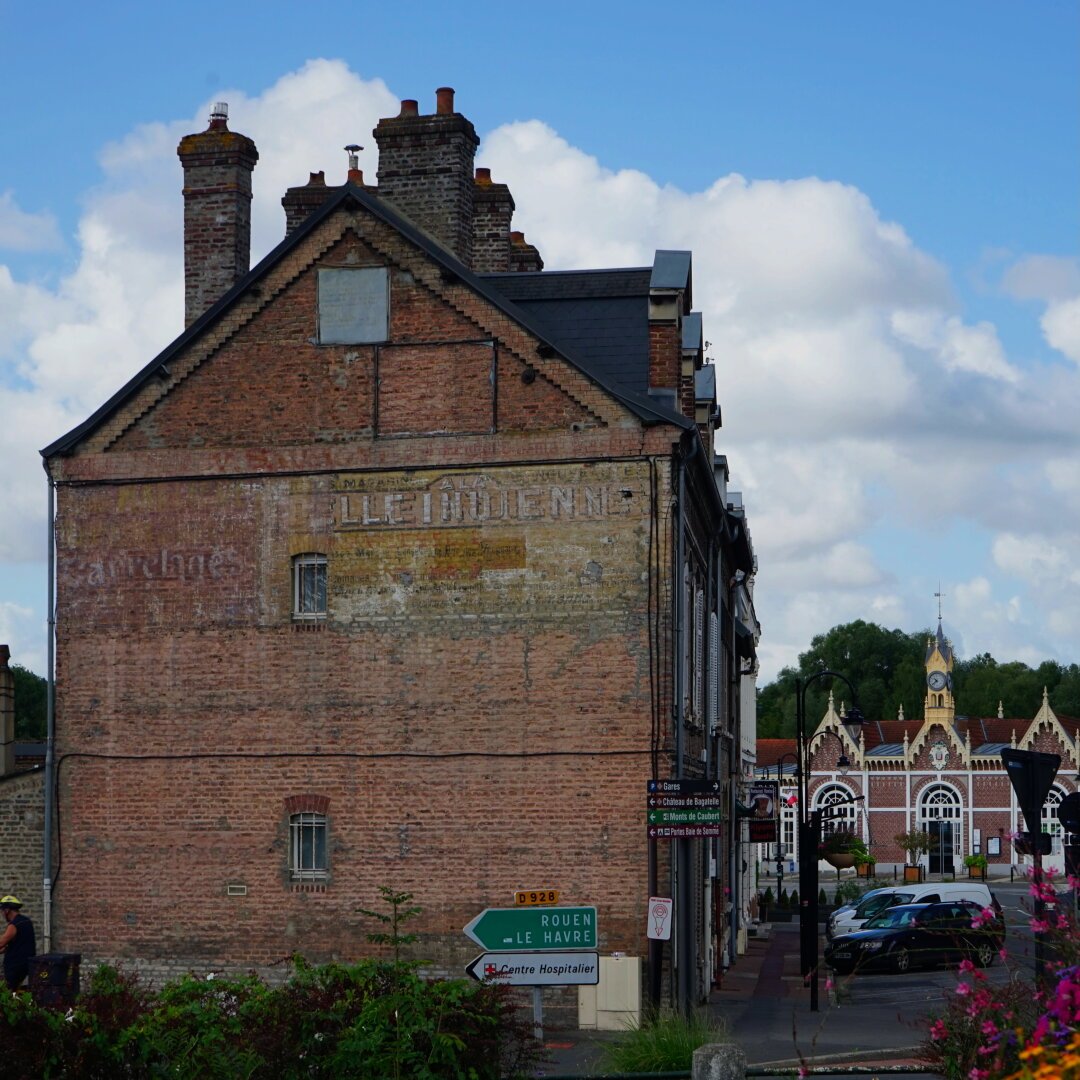 Photo montrant au fond la jolie gare d’Abbeville et au premier plan à gauche le côté d’une maison en brique assez ancienne. On y voit des traces superposées de vieilles inscriptions publicitaires à moitié effacées. On peut notamment lire le mot “carrelage” ainsi que “grand magasin À la Belle Indienne”.