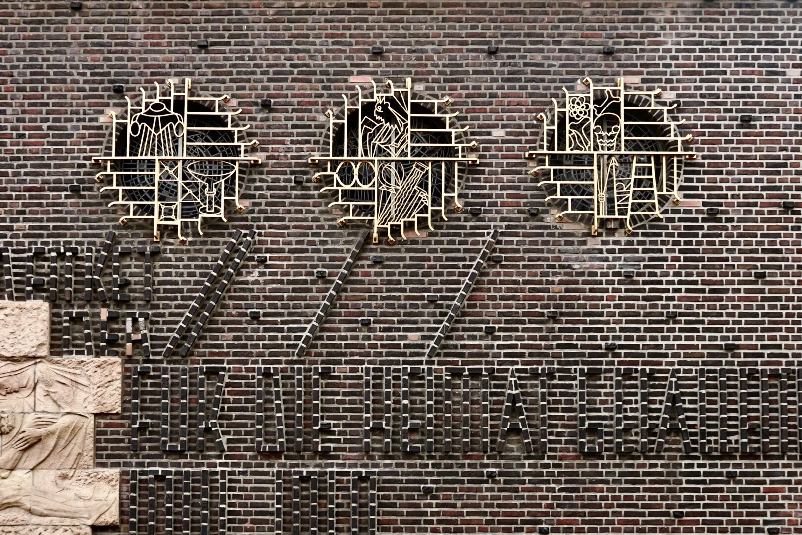 Photo d’un mur en brique marron percé dans sa partie supérieure de trois vitraux circulaires protégés par des grilles qui présentent des motifs géométriques et figuratifs (objets liturgiques, crâne et ossements). Dans la partie inférieure, on distingue difficilement une inscription en brique en relief sur le mur de briques, en lettres anguleuses et étroites (Gedenket der für die Heimat gefallenen 1914-1918). En bas à gauche, on voit l’amorce d’une décoration sculptée en pierre rose.