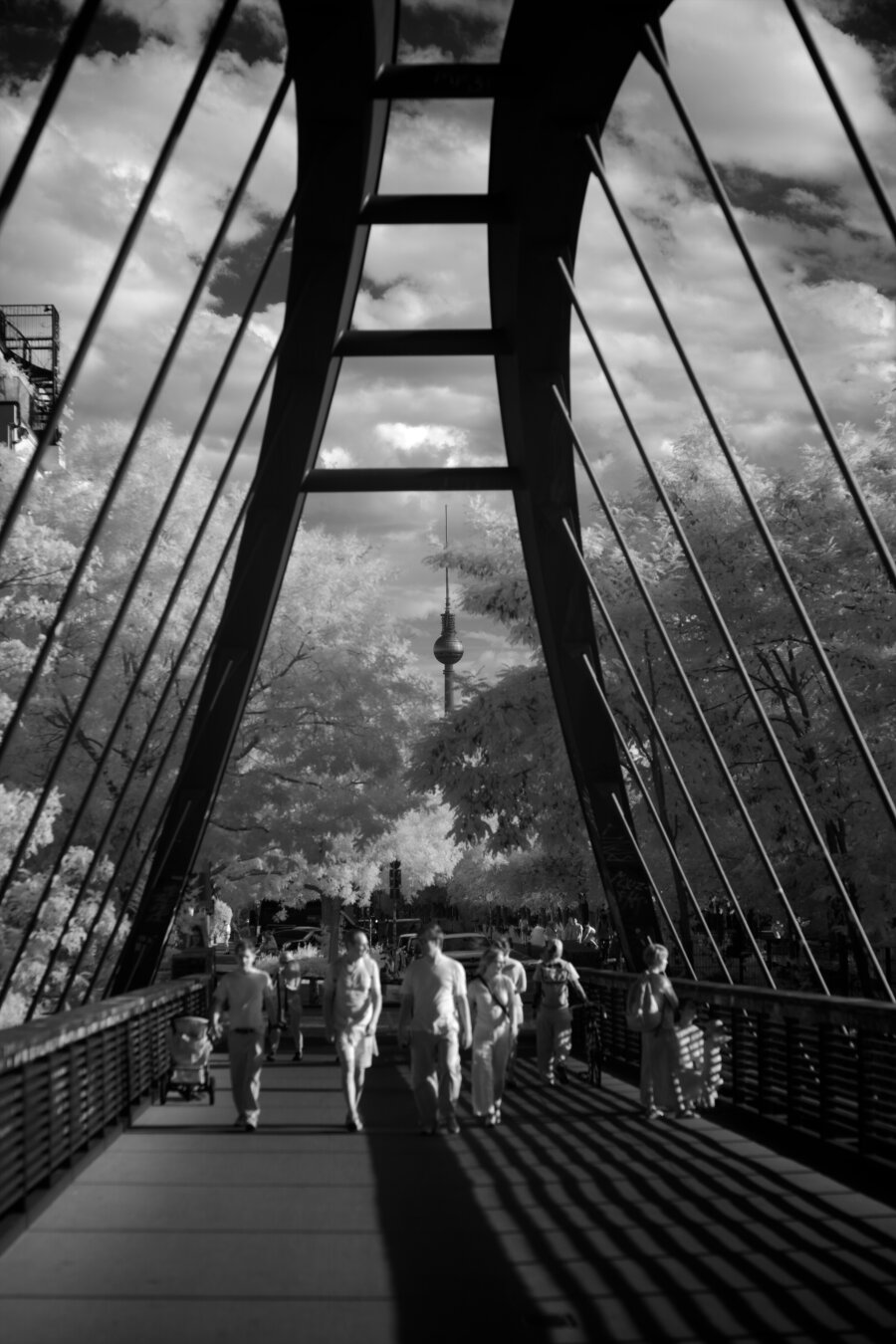 Black and white, infrared photography. The Berlin TV tower in the distance, people are walking towards the photographer, crossing a bridge. The tower is framed b the structure of the hanging bridge in the foreground.