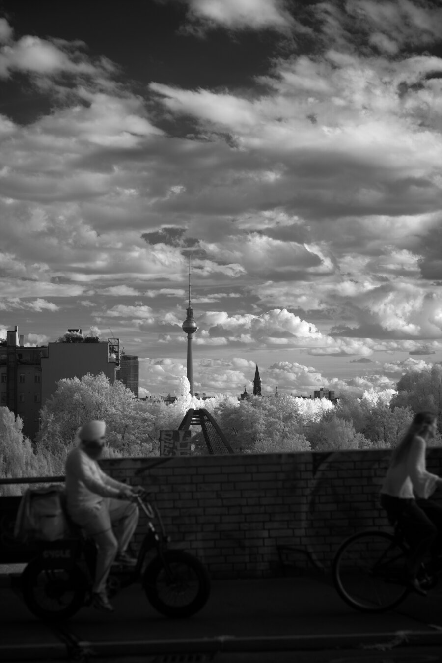 Black and white, infrared photography. The Berlin TV tower in the distance against clouds in the sky. Two people on bikes in the foreground are riding from the left to the right of the frame.