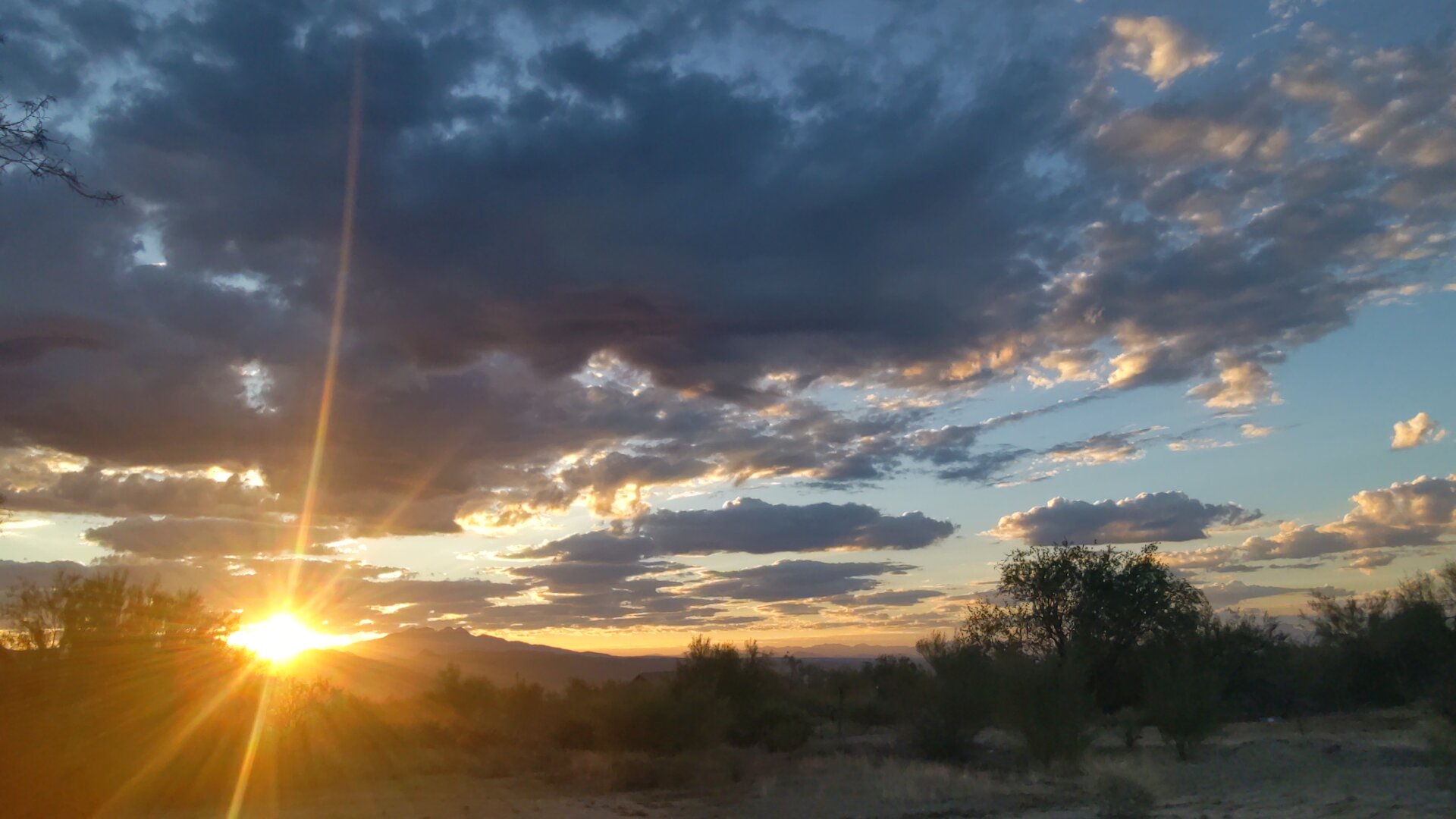 A dramatic sunrise over the Four Peaks mountain range in Arizona, with bright golden sunlight bursting through clouds and creating visible sun rays against a blue and orange sky. The silhouetted mountains and desert vegetation are backlit by the intense morning light.