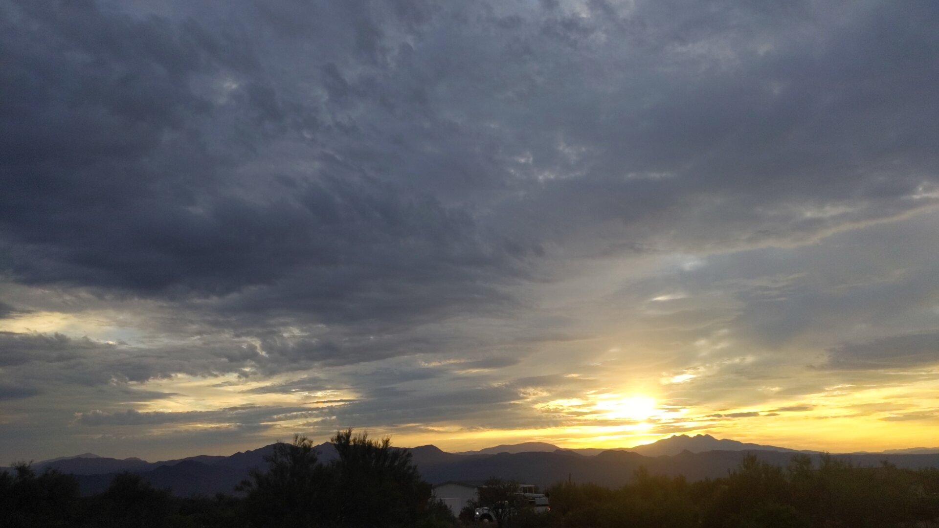 Sunrise over the Four Peaks mountain range in Arizona, with dramatic storm clouds filling the upper portion of the sky in dark blue and gray tones. Golden sunlight breaks through along the horizon, illuminating the mountain silhouettes. Desert vegetation and buildings are visible in the dark foreground.