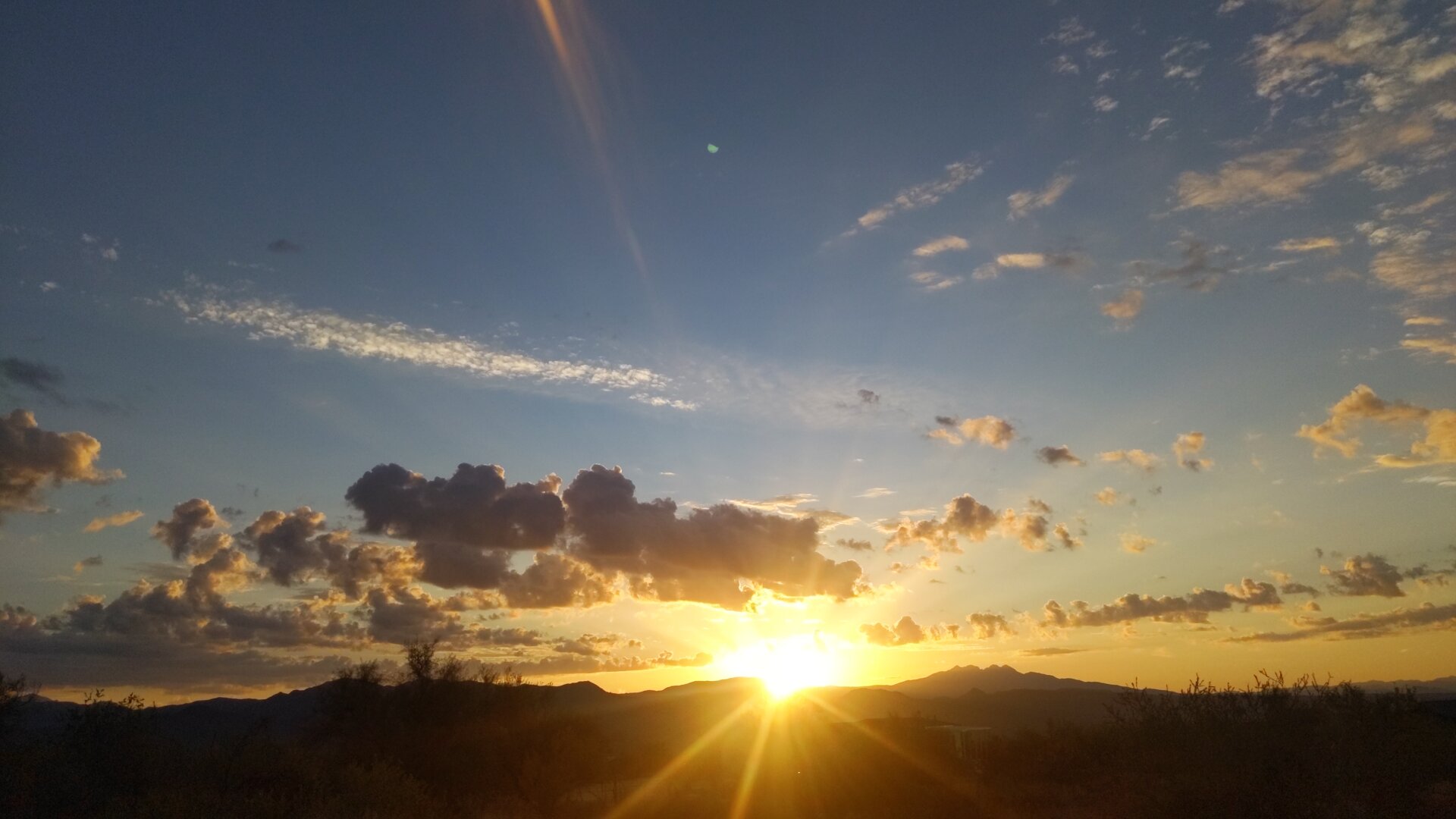 A dramatic sunrise over the Four Peaks mountain range in Arizona, with bright golden sunlight bursting through clouds and creating visible sun rays against a blue and orange sky. The silhouetted mountains and desert vegetation are backlit by the intense morning light.
