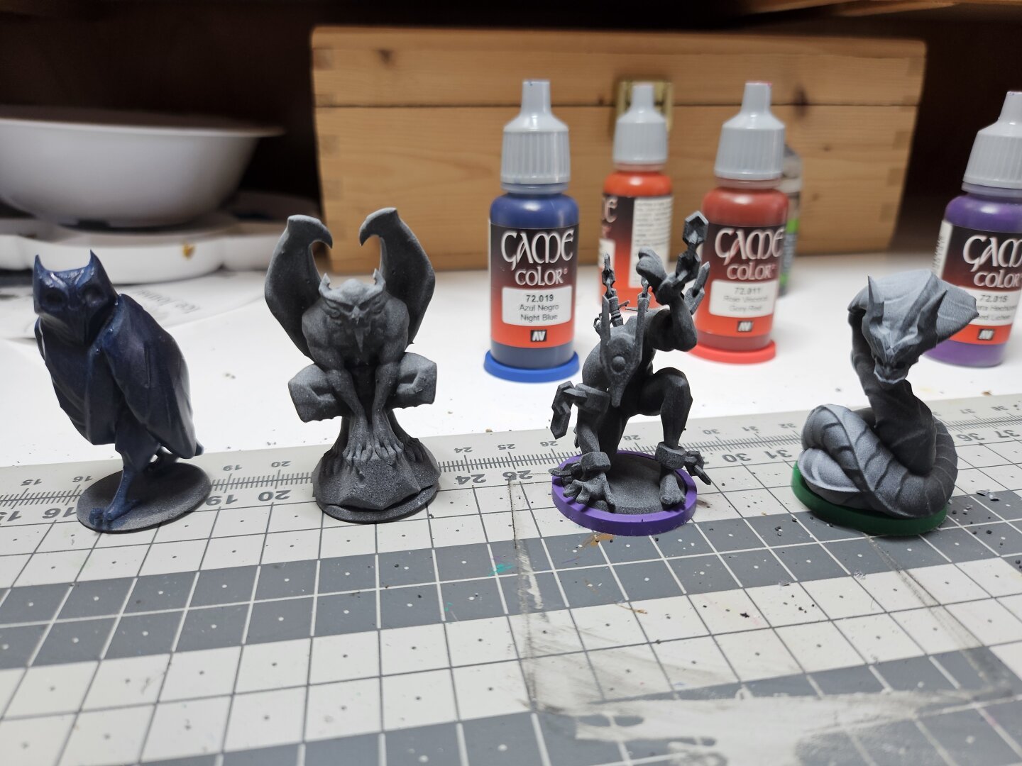 4 Minatures standing in a row. From left to right: a blueish owl, an unpainted demon perched on top of stone, a stone golem thing and a big snake.
They were part of an extra pledge for the boardgame library of jakkarth.
In the back are some game colors visible.