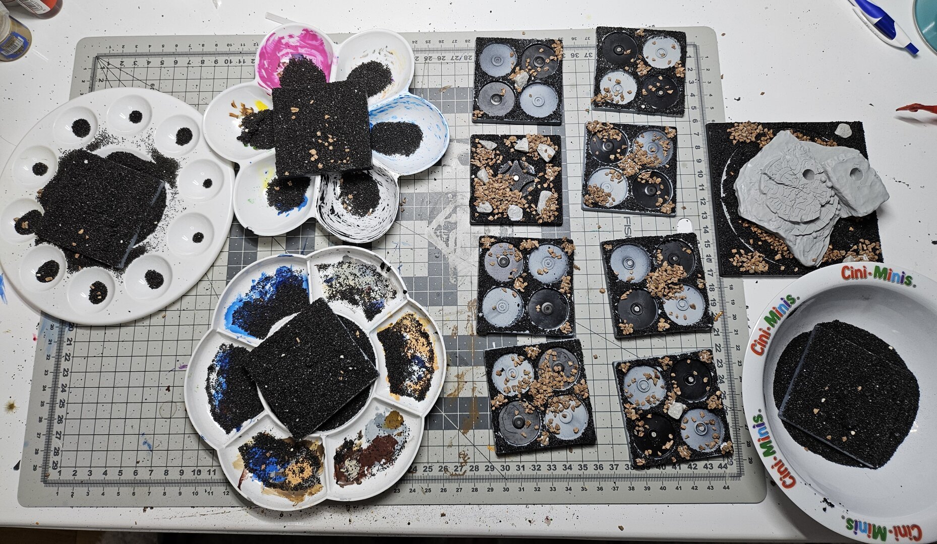 From left to right: three paint-mix-plates with conquest stands on them covered with black sand. Nine stands getting based with deko stones. One cini minis bowl containing another stand covered in black sand.