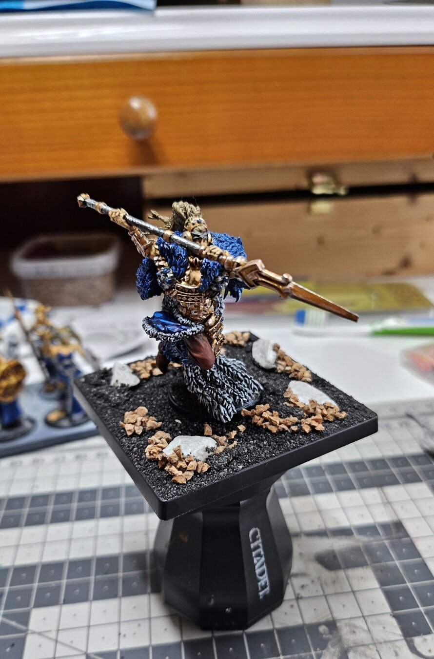 Side View of Artisan Version of Jarl of Conquest painted in blue, grey and brown, standing on an unpainted base filled with sand, tiny stones and stones.