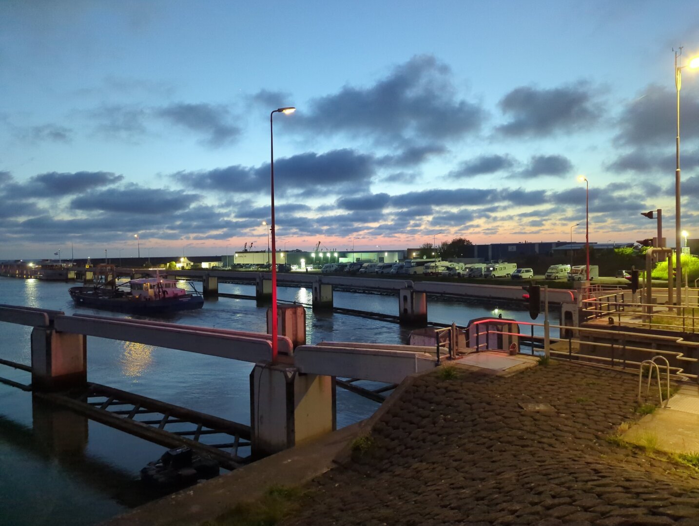 Our small white VW camper van parked at a camperstop in Harlingen haven. Left and right are big white RV's that arec3 to 4 times the sizebof ours. It's evening. The sky is blue with a pink glow from the sun setting. Some friendly clouds decorate the sky. In front there's a ship that just passed the lock, heading for sea.
