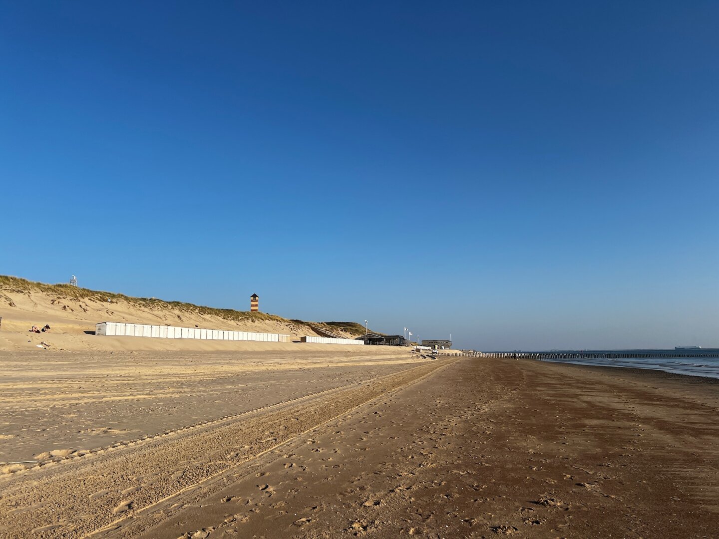 Landschapsfoto van een groot strand. Erboven een diepblauwe lucht. Links de duinen met ervoor een rij witte strandhuisjes.