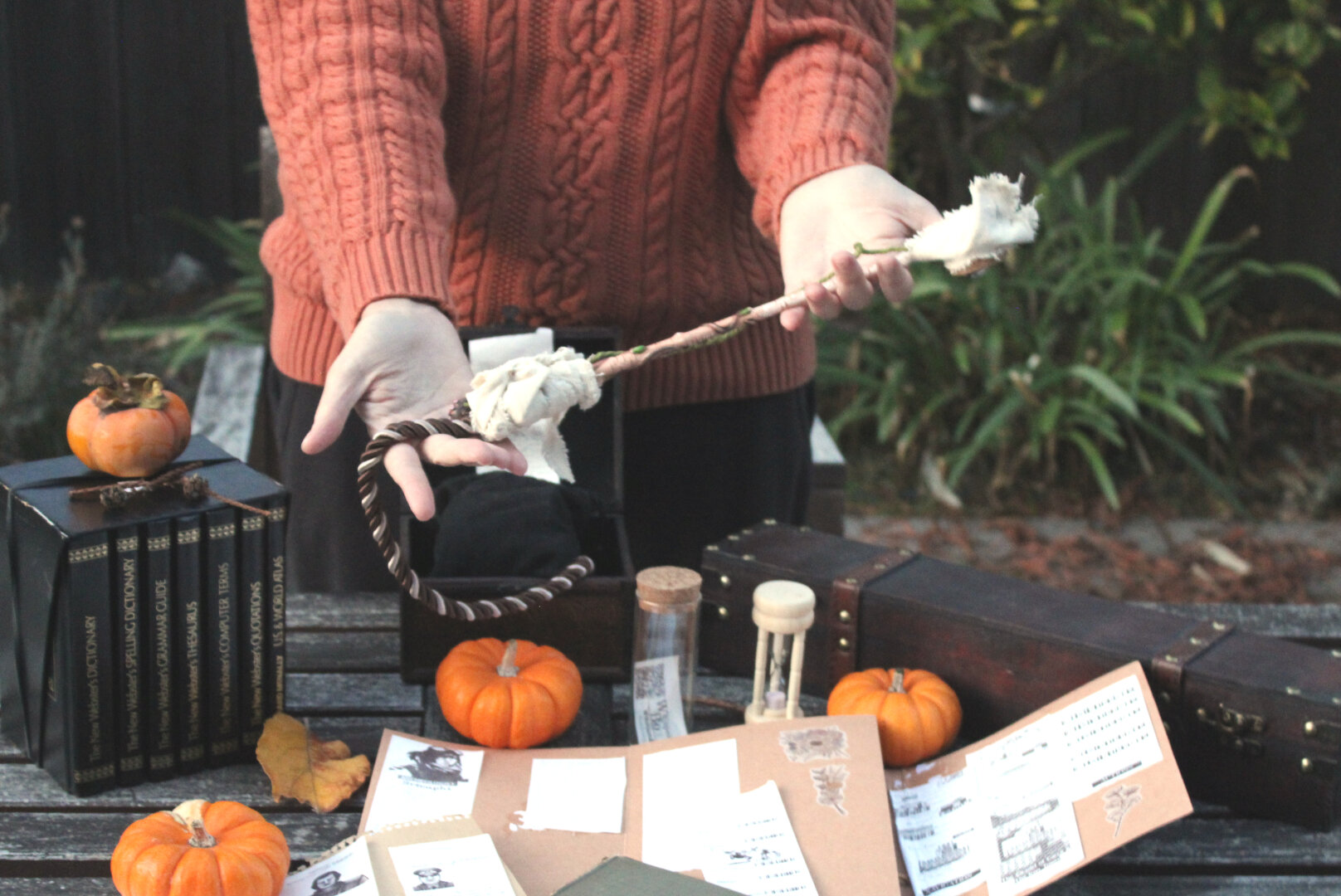 Pictured are a person facing you with their head obscured offering a wand above a wooden, outdoor desk with pumpkins, scrapbooks,  and ornate book sets, covered with bar codes and glass jars with scraps of paper.