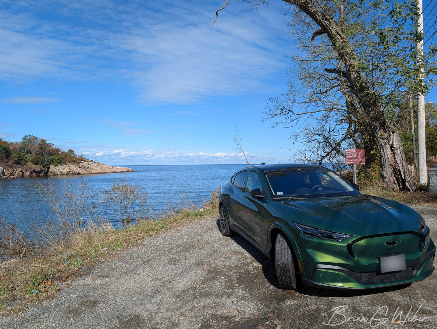 A green Ford Mach-E sits on a dirt lot in front of an ocean cove with bright sunshine, blue skies, and wispy clouds.