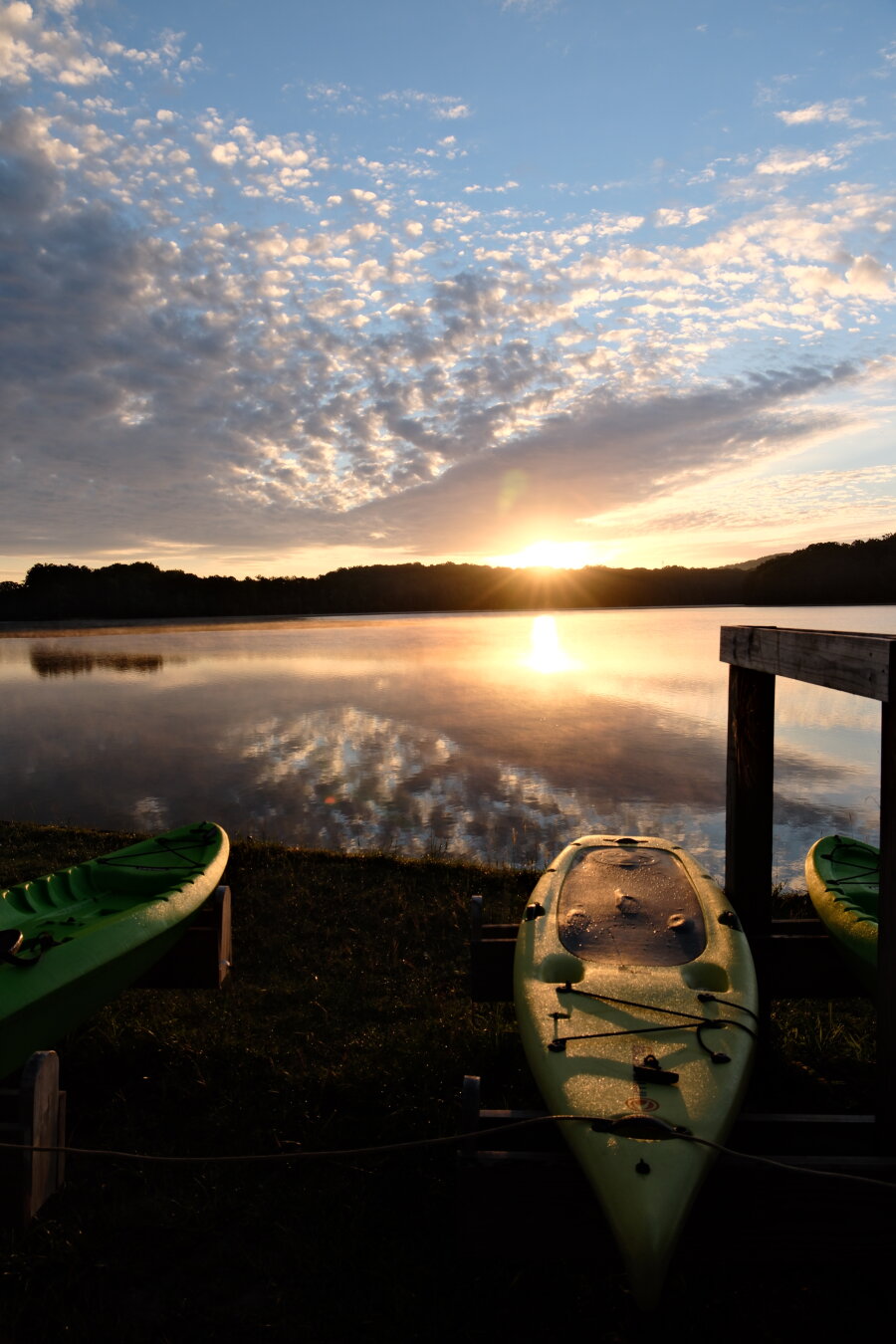 A lakeview with the rising sun. Blue sky and white clouds. Orange reflection on the water. Kayaks on the rack are in the front.