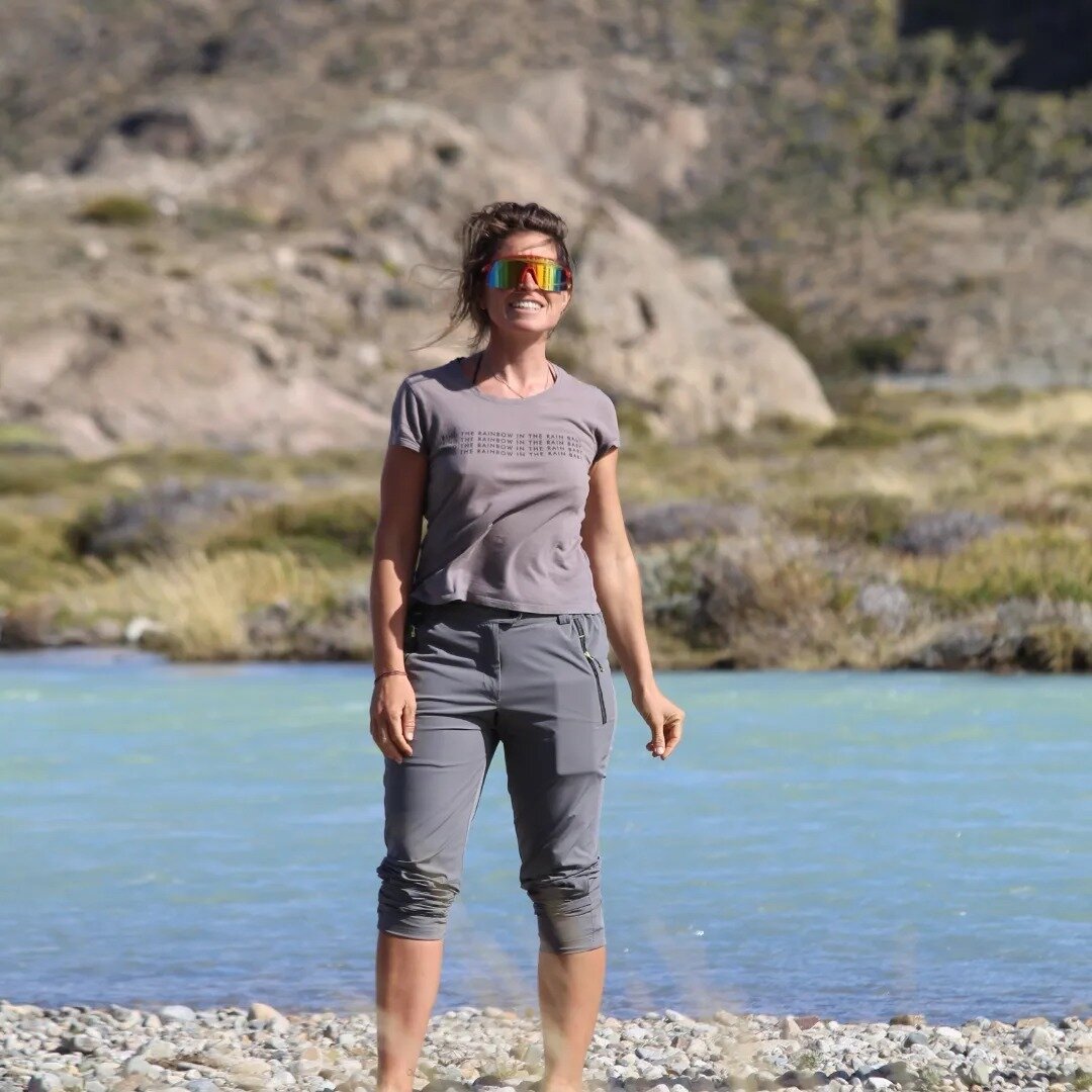 A woman wearing a grey t-shirt, grey cargo pants, and reflective sunglasses is standing on a rocky riverbank. She is smiling broadly with a backdrop of turquoise water and rugged, rocky hills.