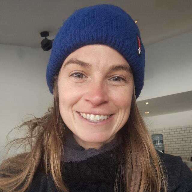 A close-up portrait of a woman smiling warmly. She is wearing a blue knit beanie and a dark scarf. The background appears to be indoors.