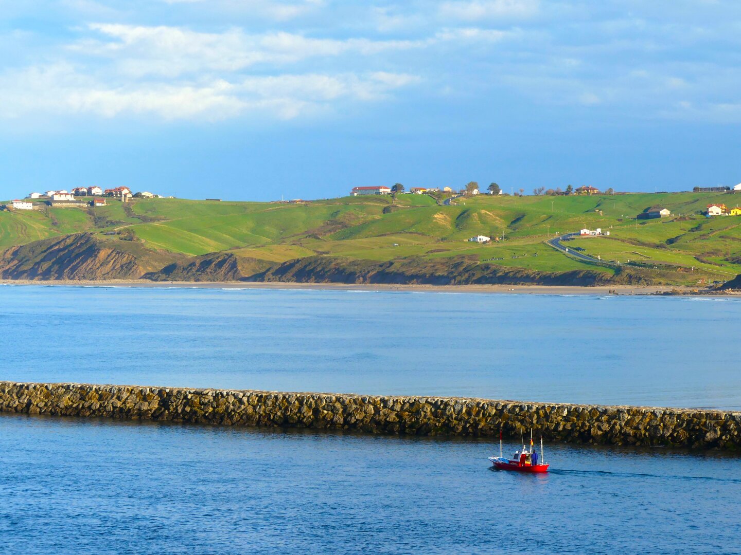 A small red boat next to a breakwater heading out to sea, with a green landscape and cliffs leading down into the water in the background.