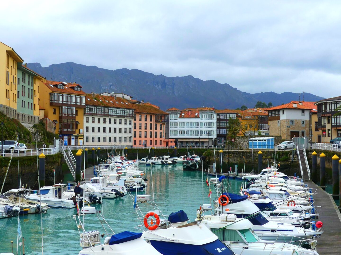 Some colorful 3- and 4-story apartment buildings above a greenish-colored inlet from the sea with many docked boats on either side, and some mountains in the background with a gray sky above.