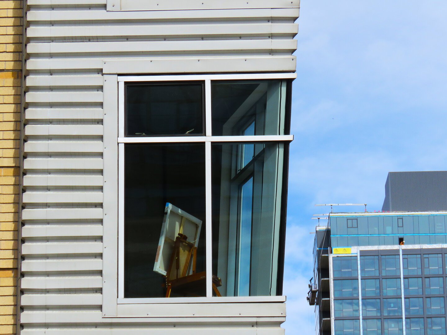 Photograph showing a corner window in a high rise residential building in Denver, Colorado, USA. In the window facing the viewer is a canvas painting which presumably someone has been working on since it sits on an easel, though there are no people visible and the easel could just be for display. In the background is another high rise building.