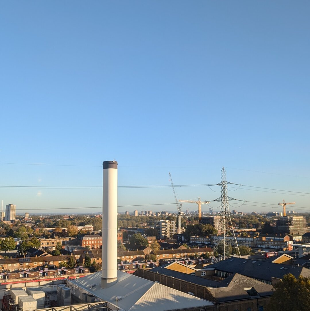 A view north from London's docklands, from a high floor. A big white chimney is surrounded by much shorter beige brick buildings, concrete office blocks and pylons, all under bright clear blue sky.