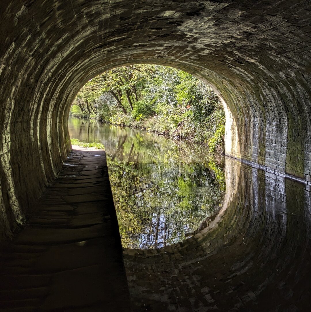 View along a canal tunnel toward sunlit greenery. The towpath to one side of the canal, the arched stone tunnel above, and their reflections make a snail shell shape.