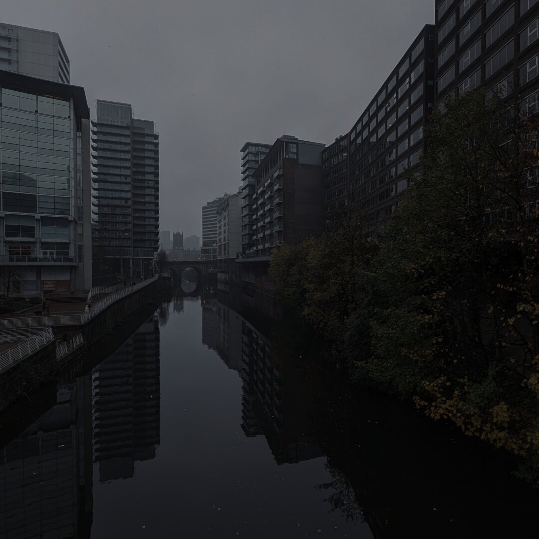 View North up the River Irwell on a rainy day, taken from Trinity Bridge. Tall buildings recede into the gloom. In the distance, the spire of Manchester Cathedral.