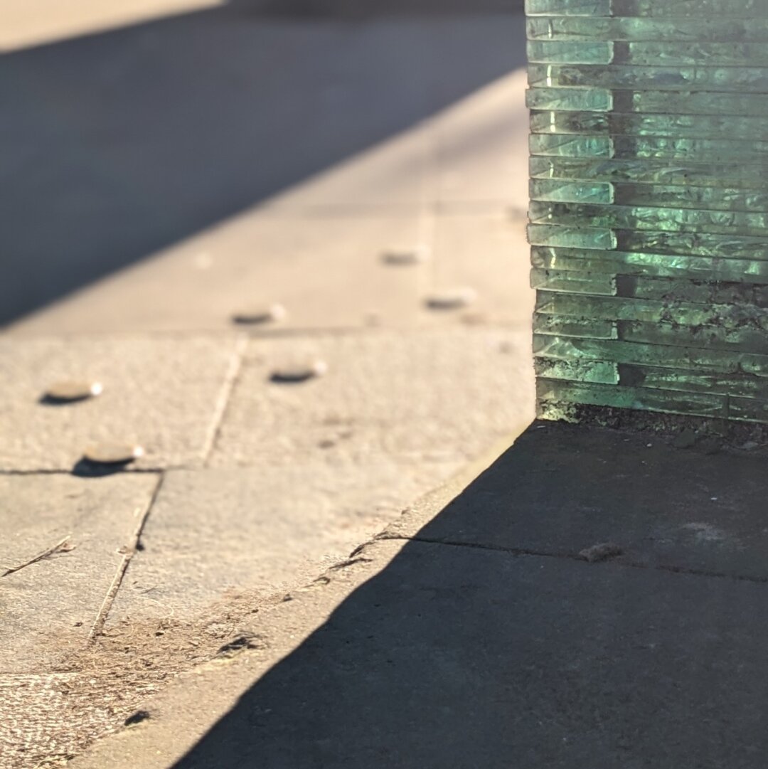 Close up shot of a bench made from stacked glass sheets, with bright winter sun streaking through them. The glass shines bright blue, and the sun draws sharp long shadows over the brownish flagstones beneath