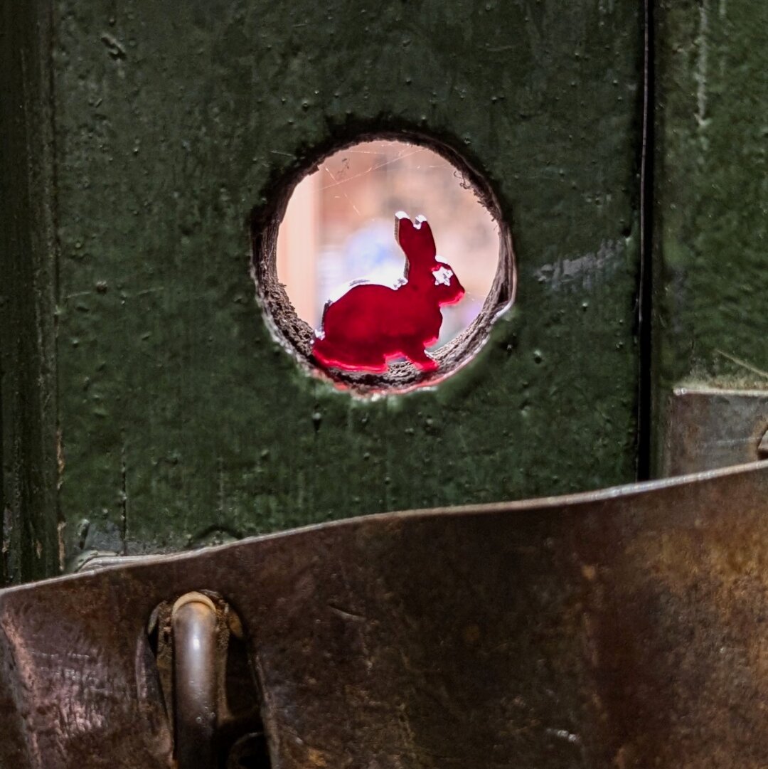 Closeup of a green wooden door, with a hasp that's closed but has no padlock. Above it, a small hole through the door has a small, red laser cut rabbit sitting in it.