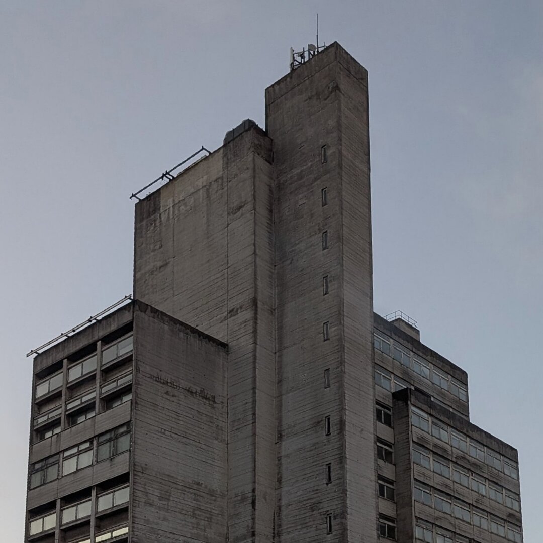 A very grey concrete high rise building, a cacophony of stacked oblongs, near twilight against pale blue sky.