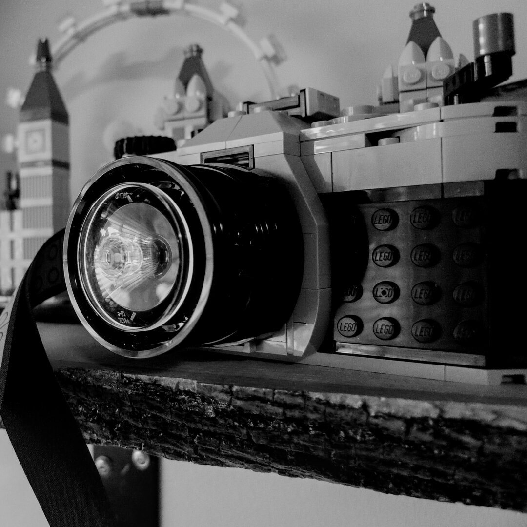 Black and white photo. On the right, in the foreground is Lego camera. On the left, in the background is Big Ben (also in Lego). Both are positioned on a live edge wood shelf.
