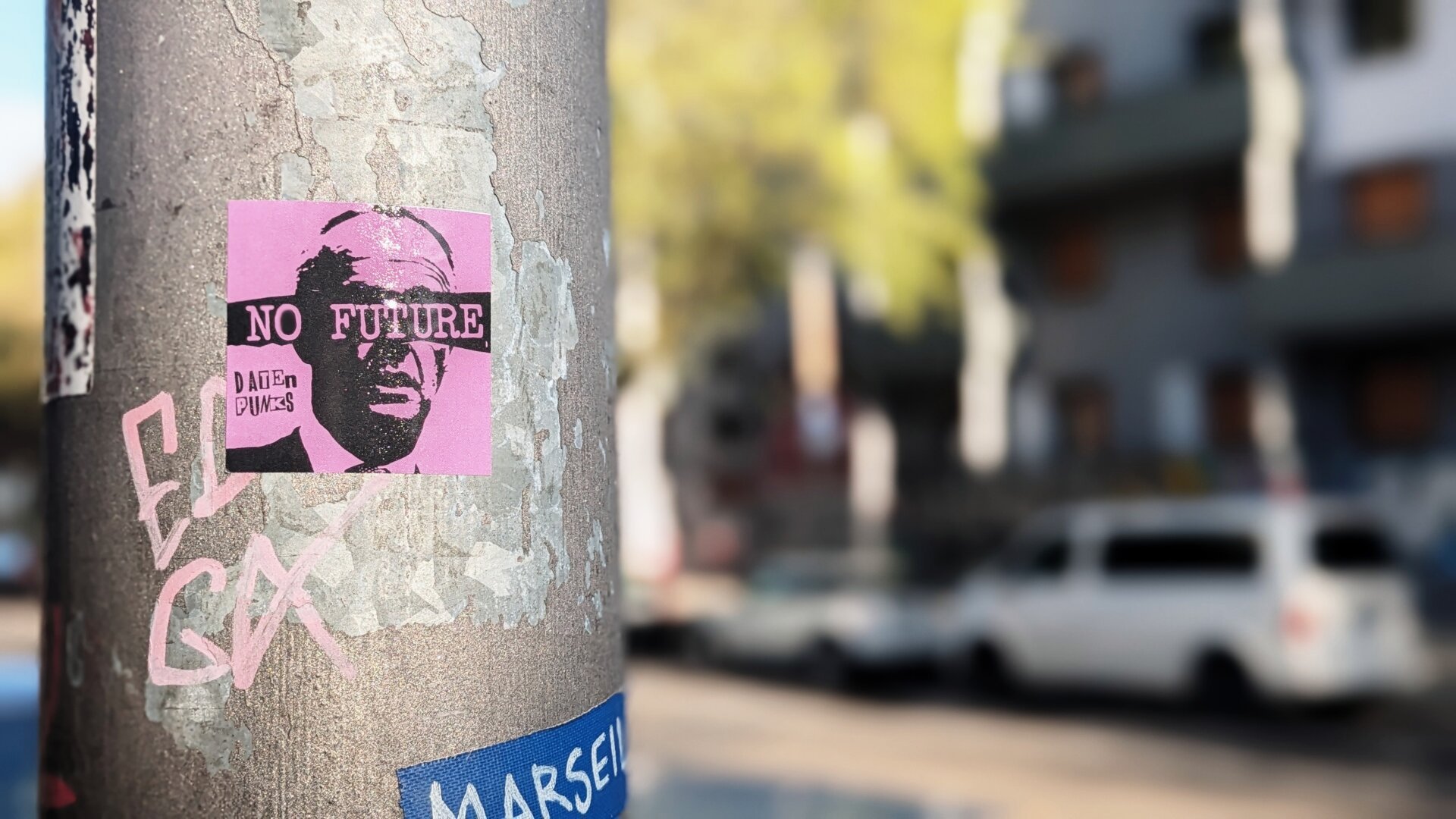 Der Pfosten einer Strassenlaterne. Darauf ein violetter, quadratischer Aufkleber mit einem Bild von Friedrich Merz und der Aufschrift "No Future" über den Augen.