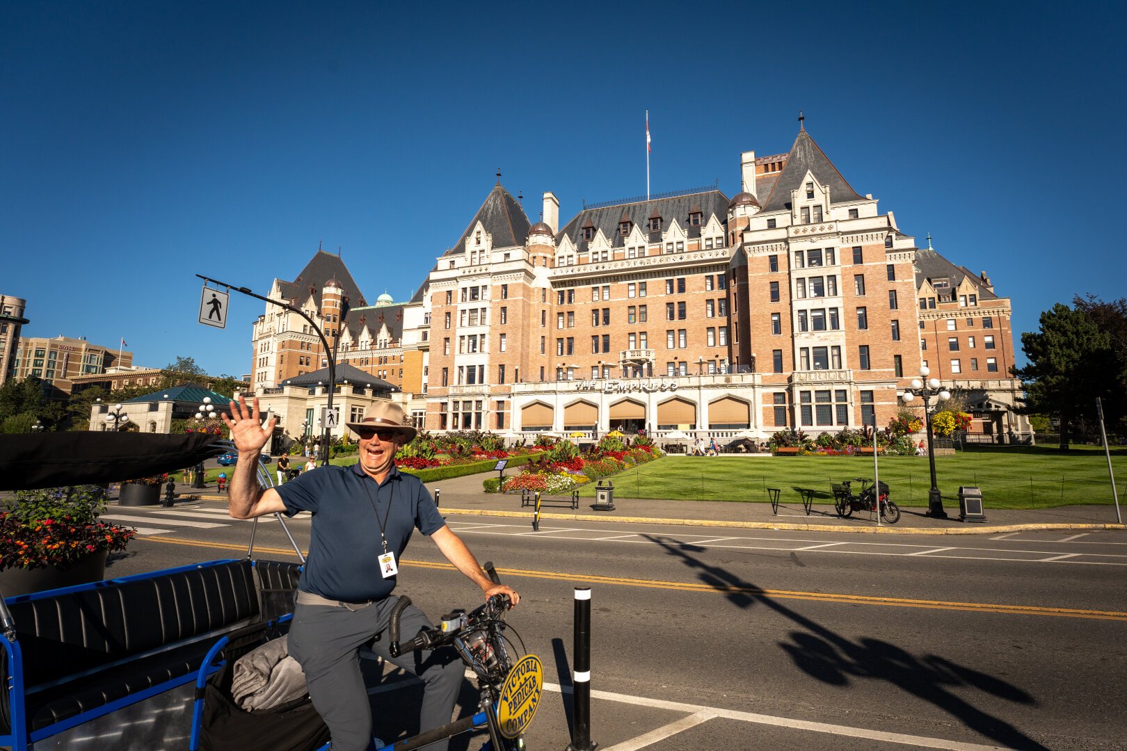 The Empress hotel, a large and fancy lookin' building sits across the street while a smiling man waves at the camera as he peddles a pedicab down the street.
