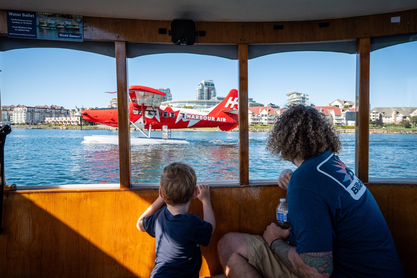 A young boy and his father look out the window of a small boat to watch a red and white seaplane with a maple leaf logo and Harbour Air insignia as it begins take off from the harbour.